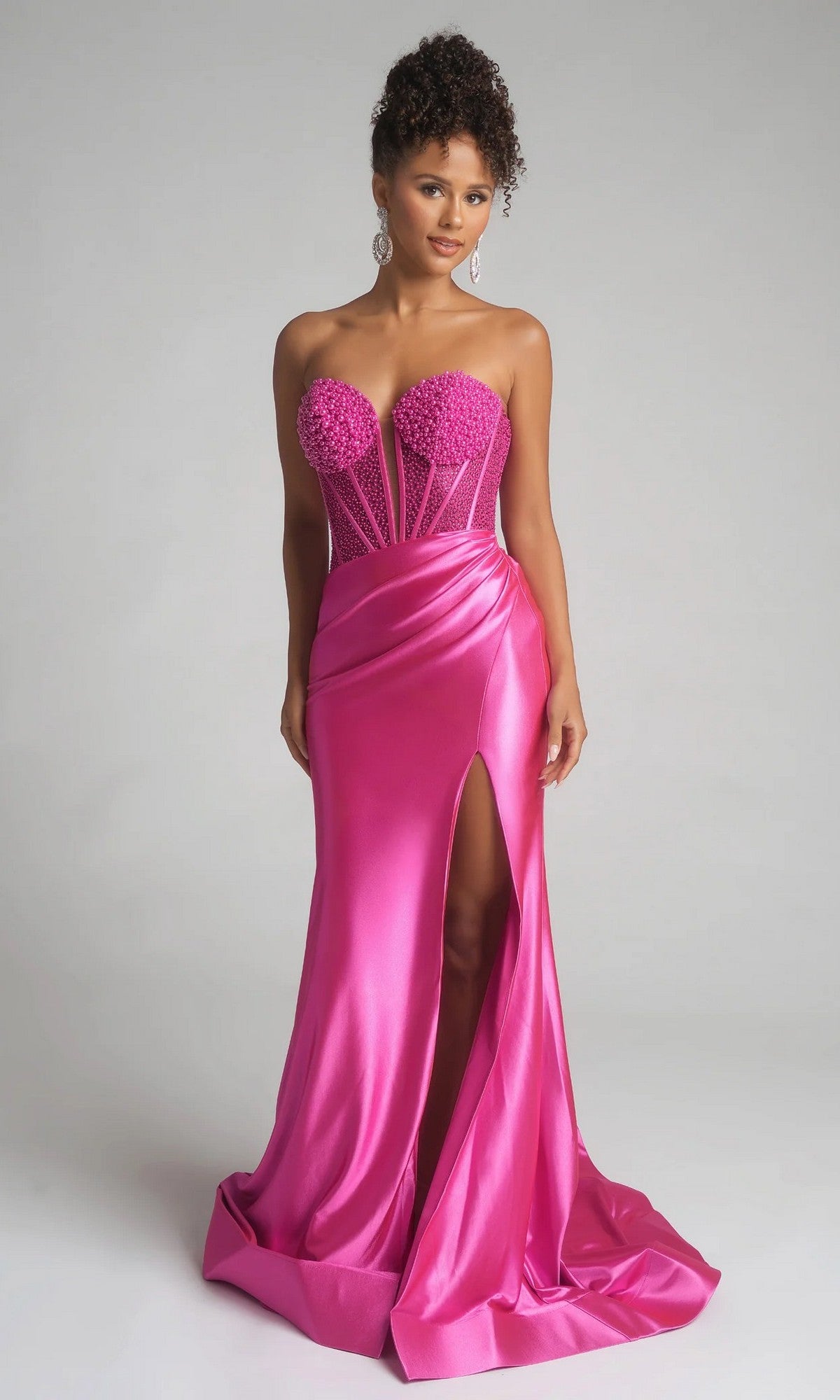 A woman stands against a plain background in the Portia and Scarlett PS26039 long prom dress in fuchsia, featuring a strapless beaded corset bodice, floral details, and a high slit.